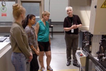 Graphic and Print Technician students standing while their instructor gives an example of using a commercial print machine.